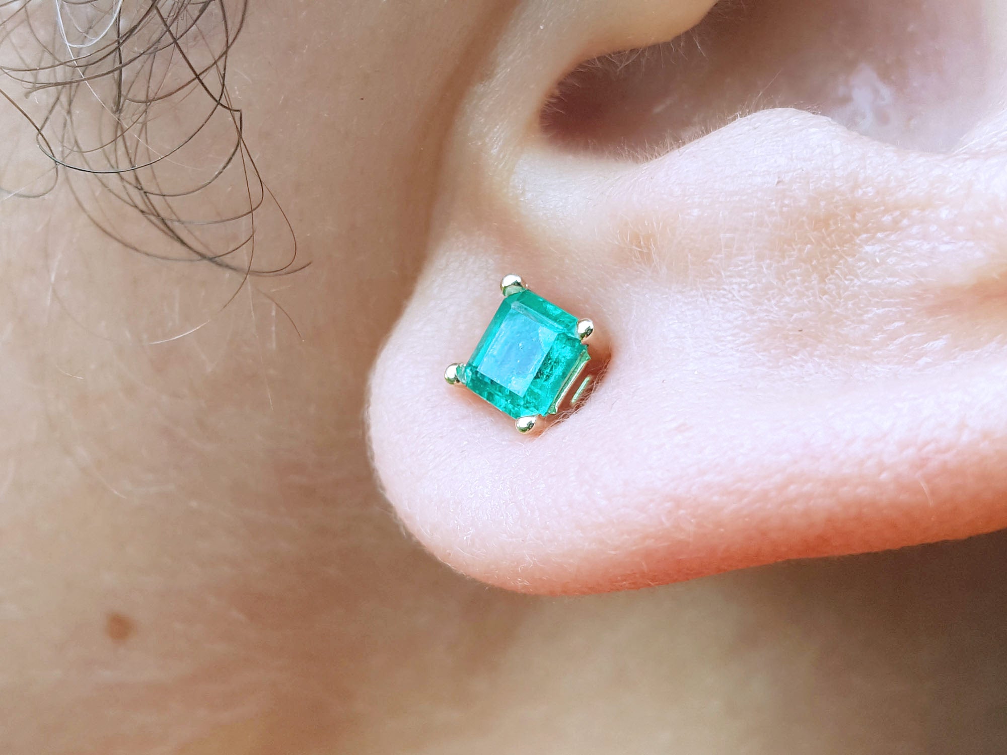 Close-up of a yellow gold square emerald stud earring on an earlobe.