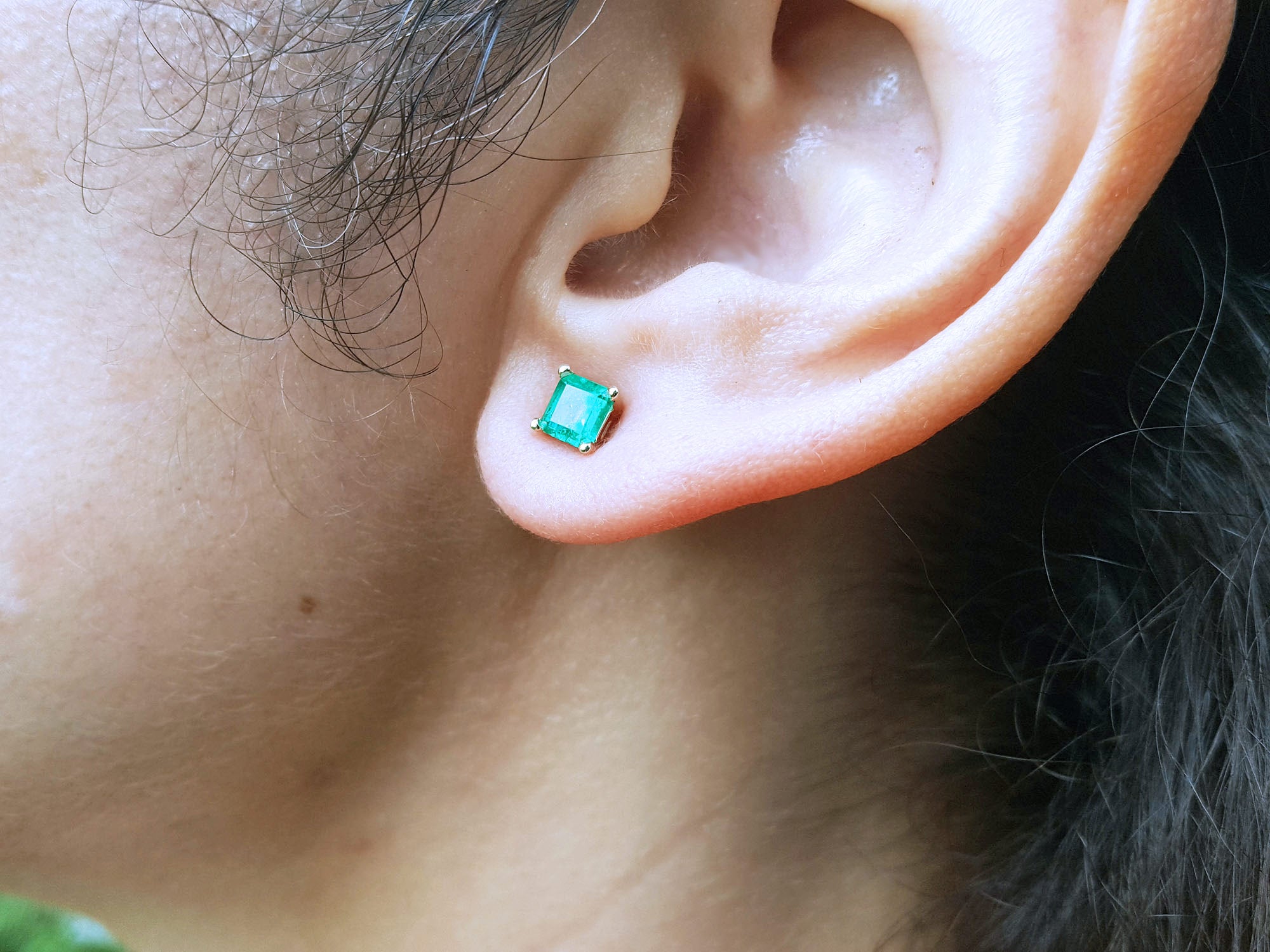 Close-up of a woman&#39;s ear wearing a square yellow gold emerald stud earring with hair and skin visible.