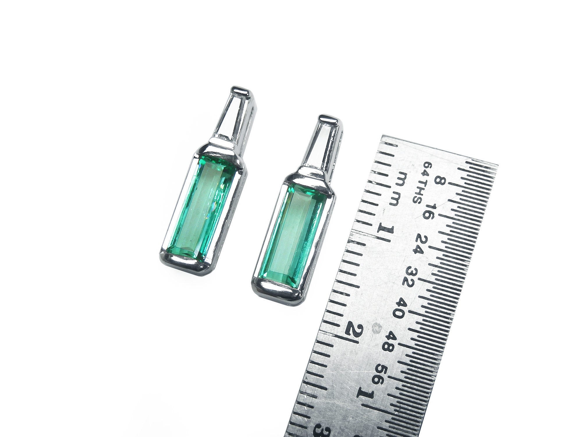 Two Muzo Colombian baguette emerald stud earrings next to a ruler for scale on a white background