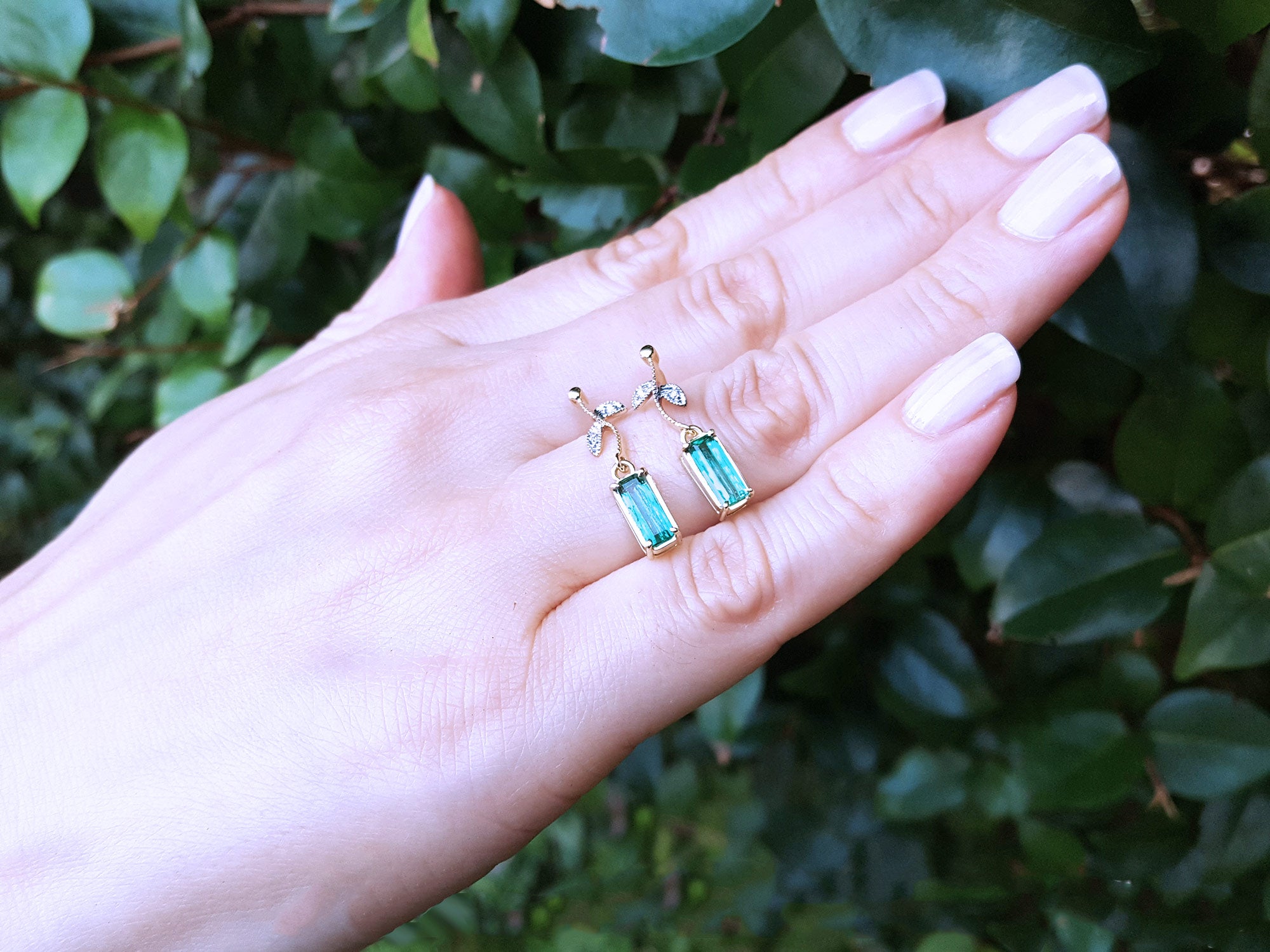 Hand displaying two emerald gemstone earrings with a green leafy background