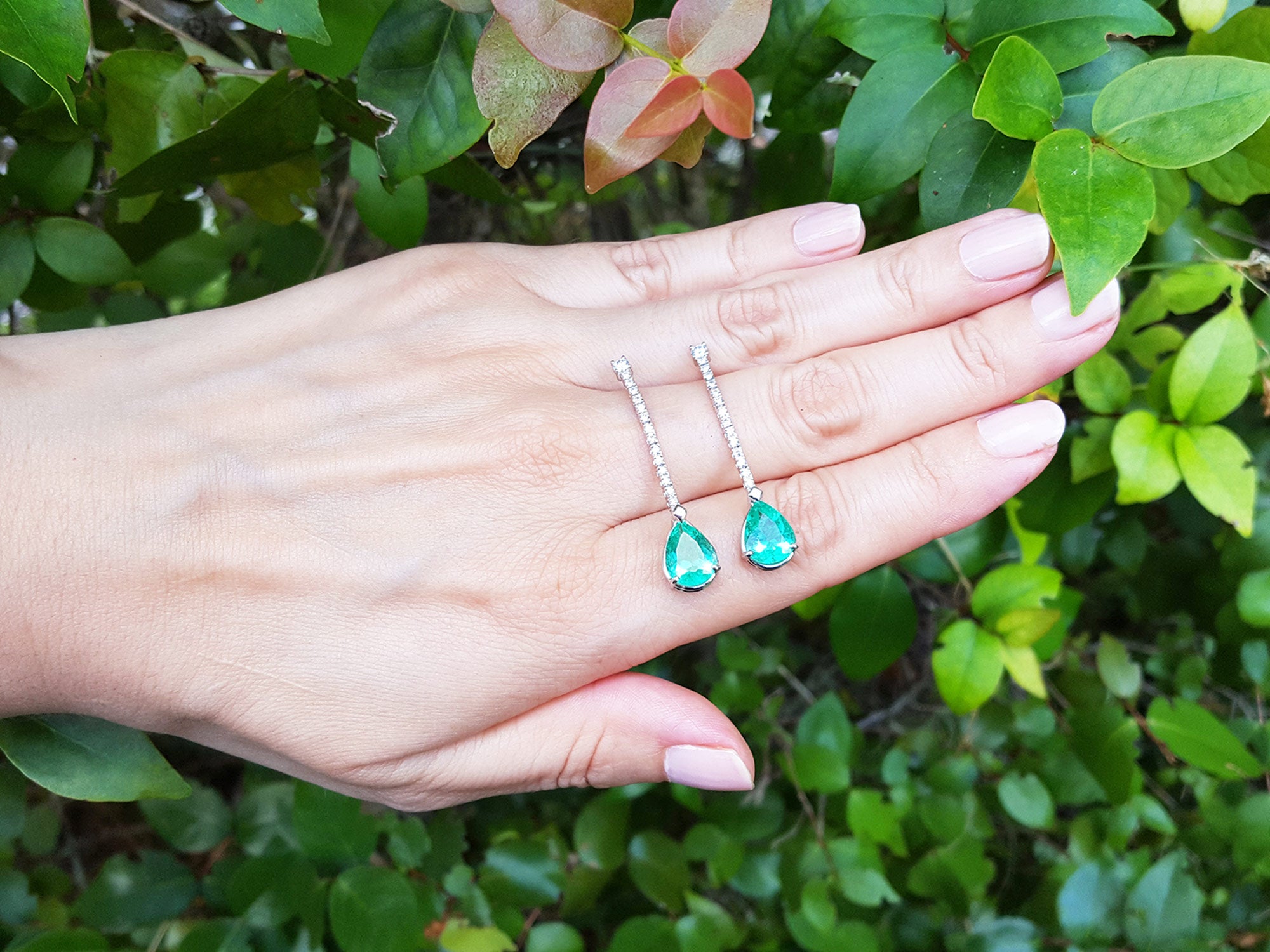 Hand holding two green teardrop earrings against a green leafy background