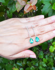 Hand holding two green teardrop earrings against a green leafy background