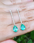 Green teardrop earrings held between fingers with a blurred natural background