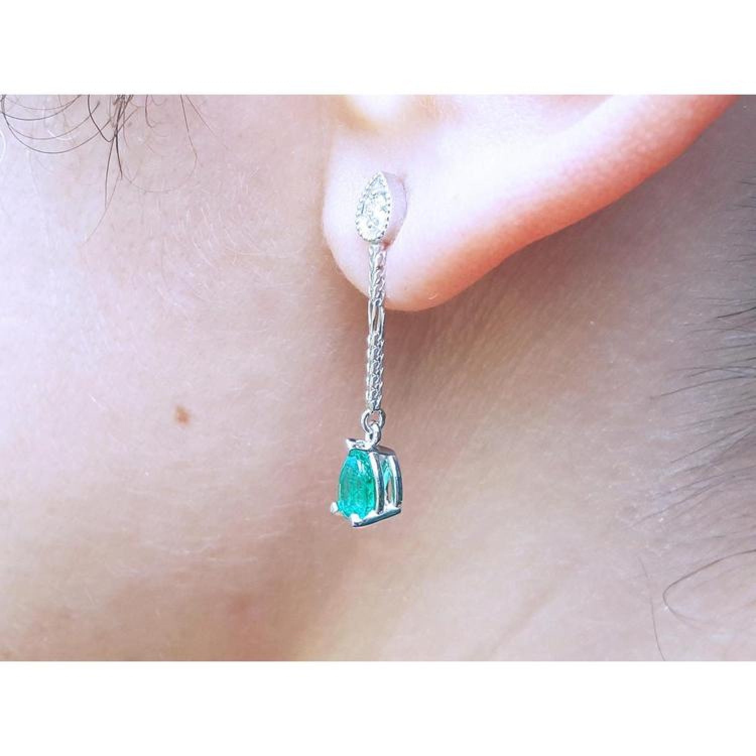 Pear-shaped Muzo Colombian emerald dangle earrings in 14K white gold with diamond accents on an ear.