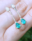 A 14k gold emerald dangle earrings placed on a hand against a blurred background
