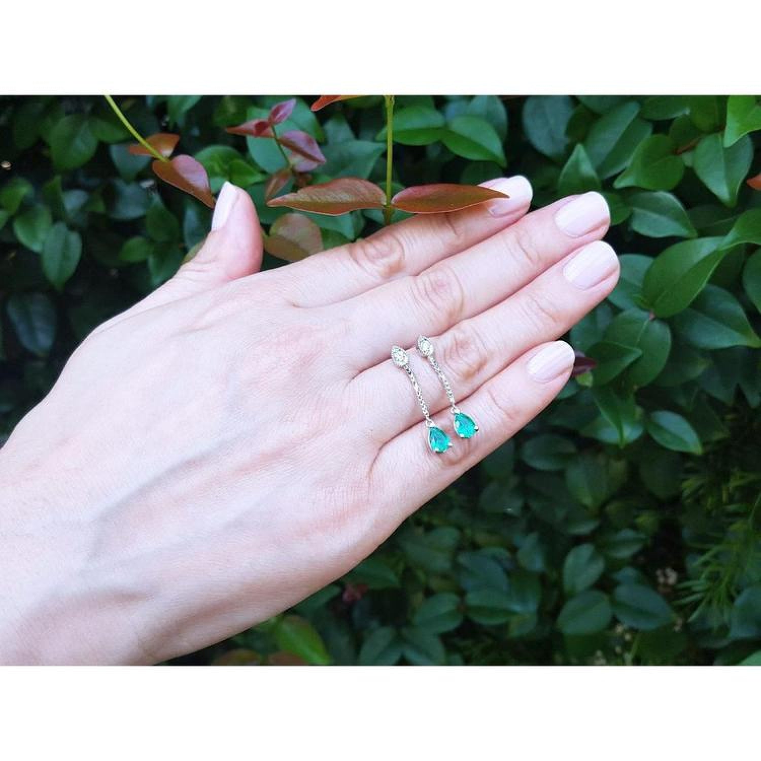Pear-shaped Muzo Colombian emerald dangle earrings in 14K white gold with diamonds, worn on a hand.