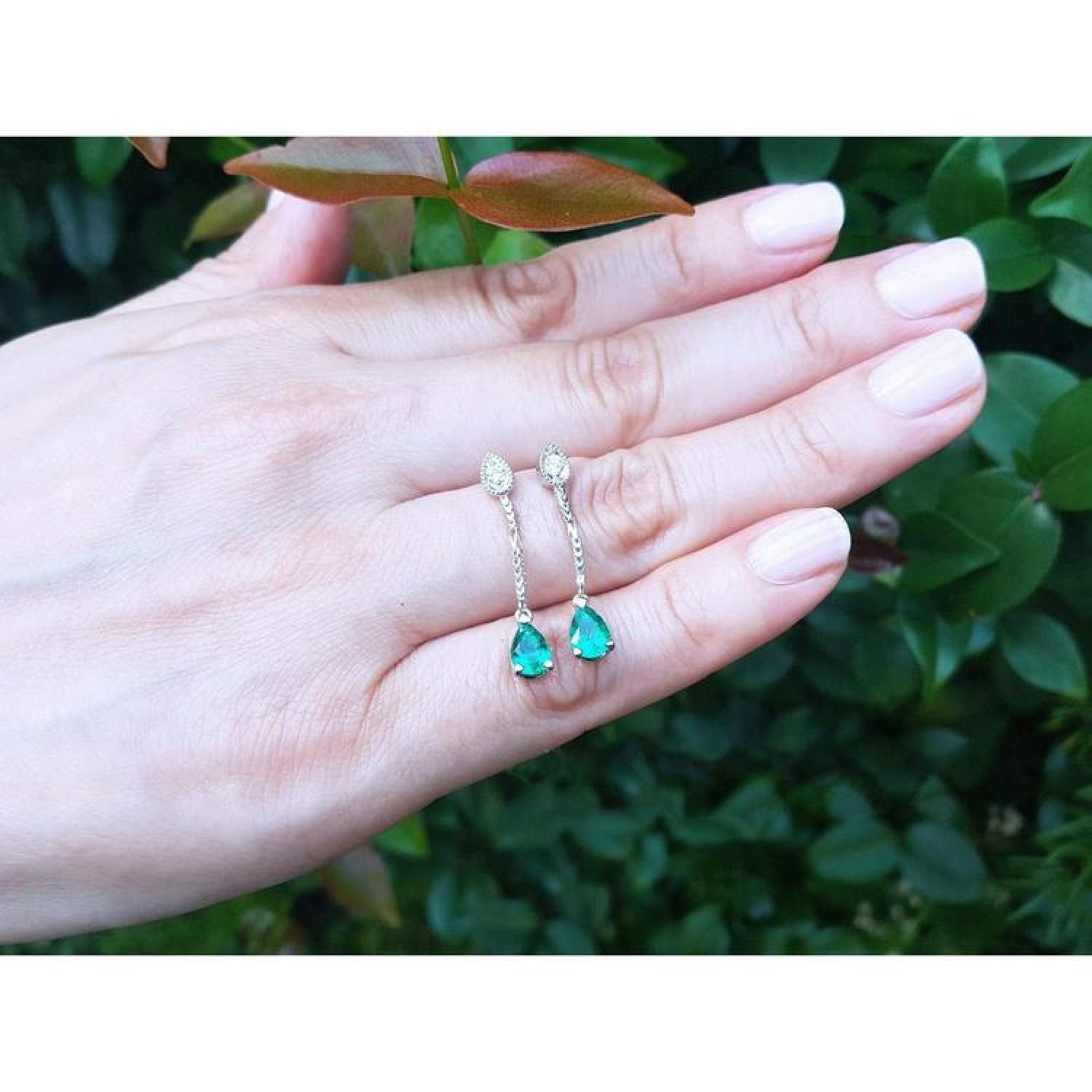Pear-shaped Muzo Colombian emerald dangle earrings with diamonds in 14K white gold on a hand.
