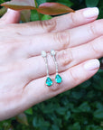 Hand displaying emerald teardrop earrings with a blurred green leafy background
