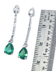 Pair of white gold emerald earrings dangle next to a ruler for scale on a white background.