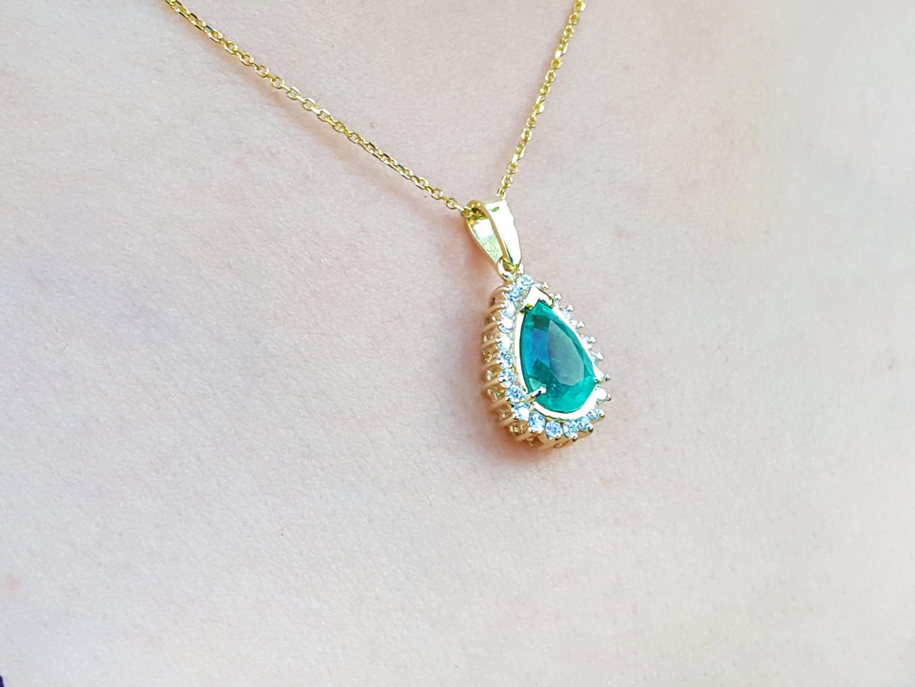 Teardrop-shaped pendant with a natural emerald on a gold chain against a light pink skin background
