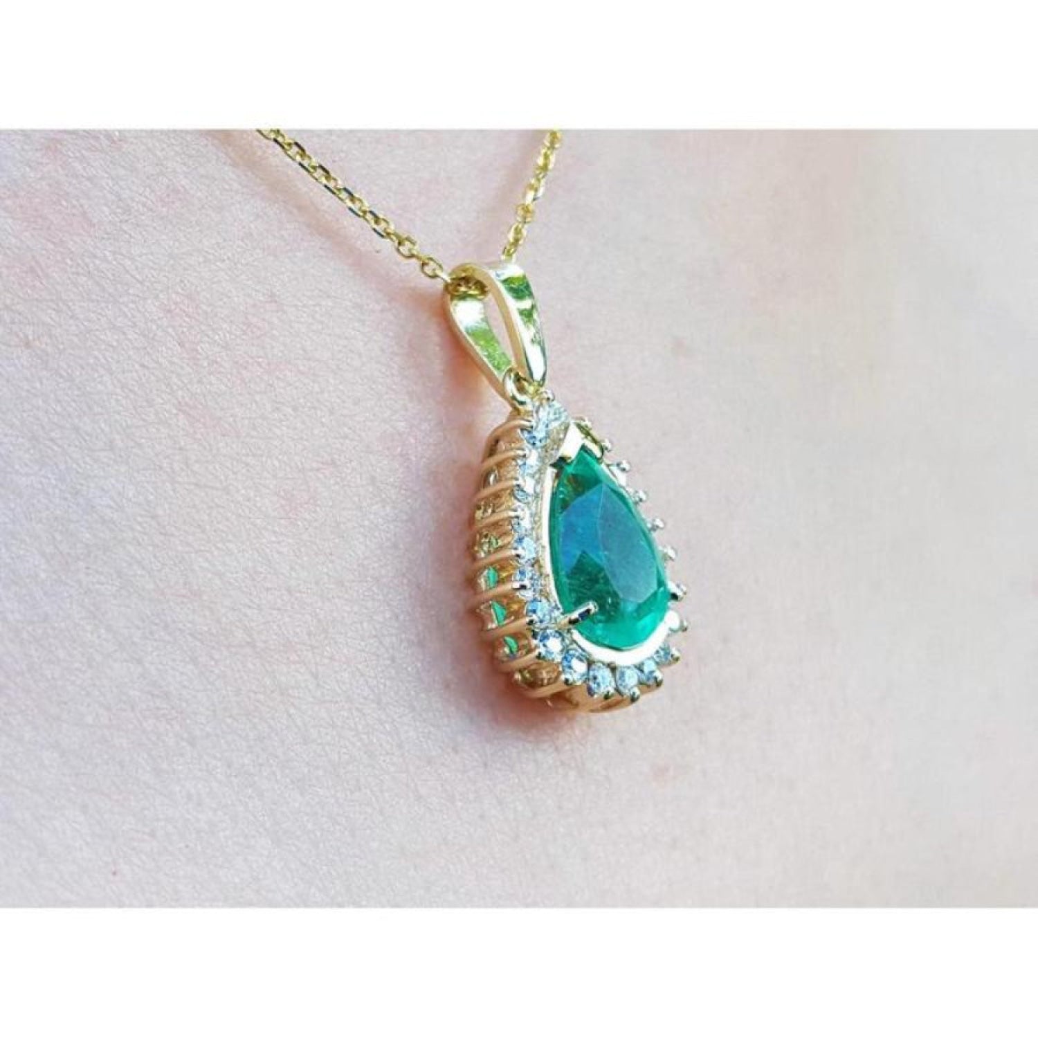 Gold pendant with a natural teardrop-shaped emerald on a light pink skin background.