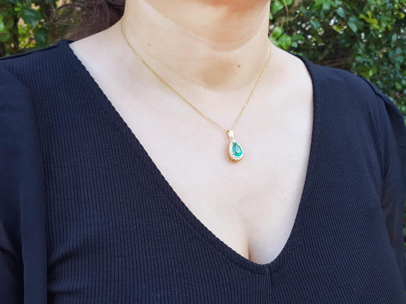 A woman wearing a gold necklace with a colombian emerald pendant against a blurred natural background