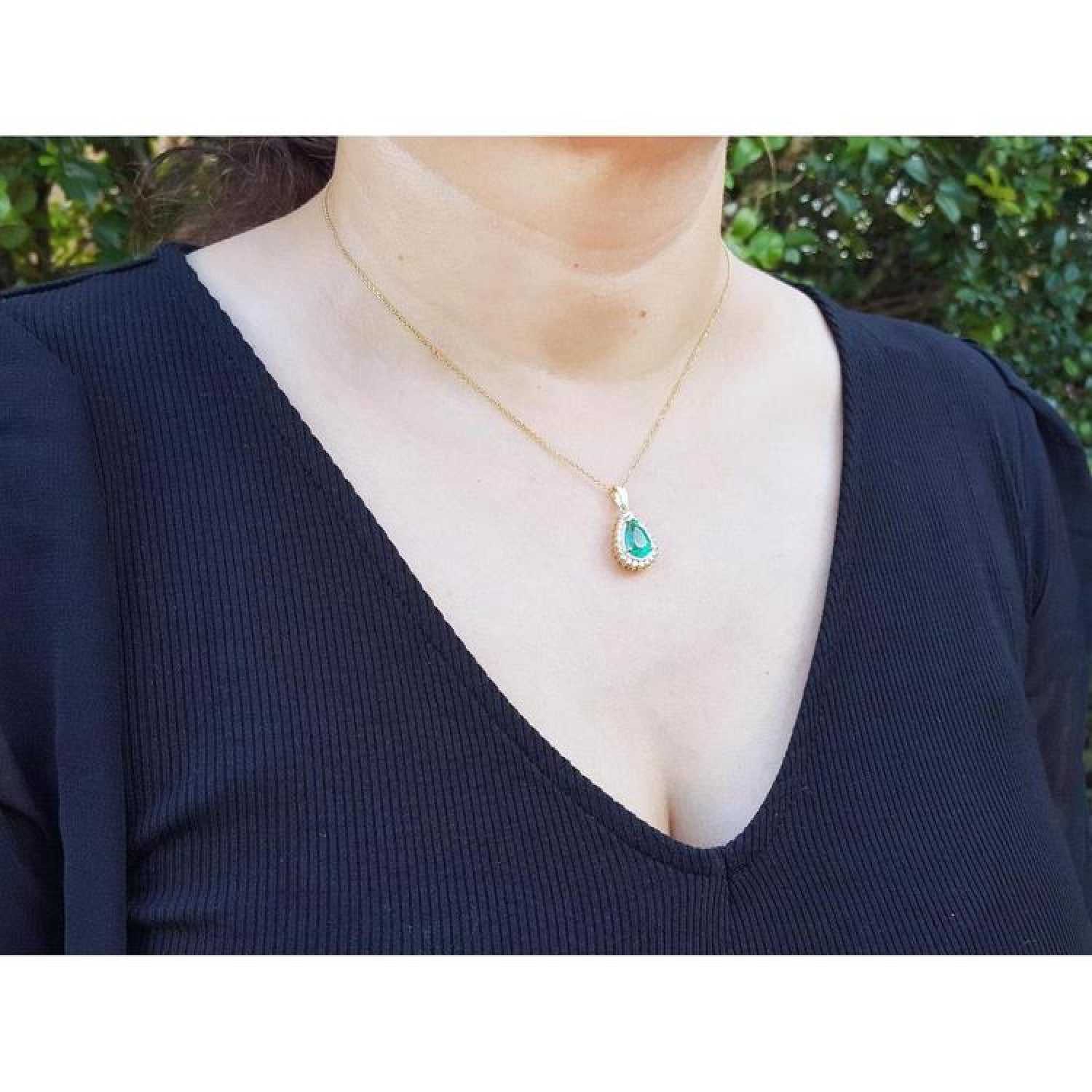 Teardrop emerald pendant with diamond halo on a gold chain, worn by a woman.