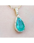 Teardrop-shaped emerald gemstone pendant with halo diamond accents on a light pink skin background