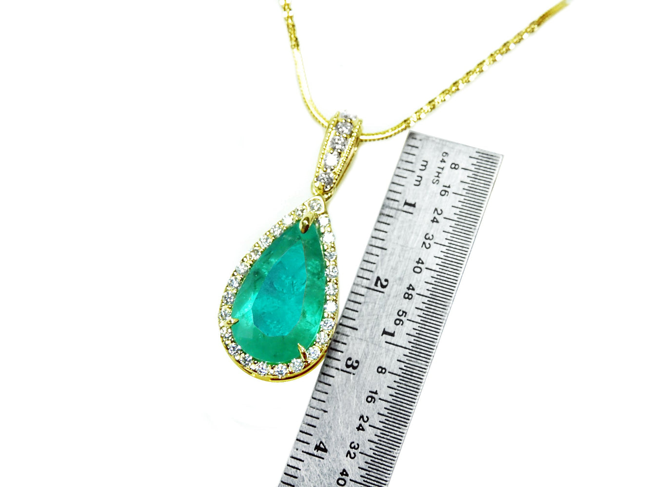 A natural colombian emerald teardrop pendant on a gold chain with a ruler for scale on a white background