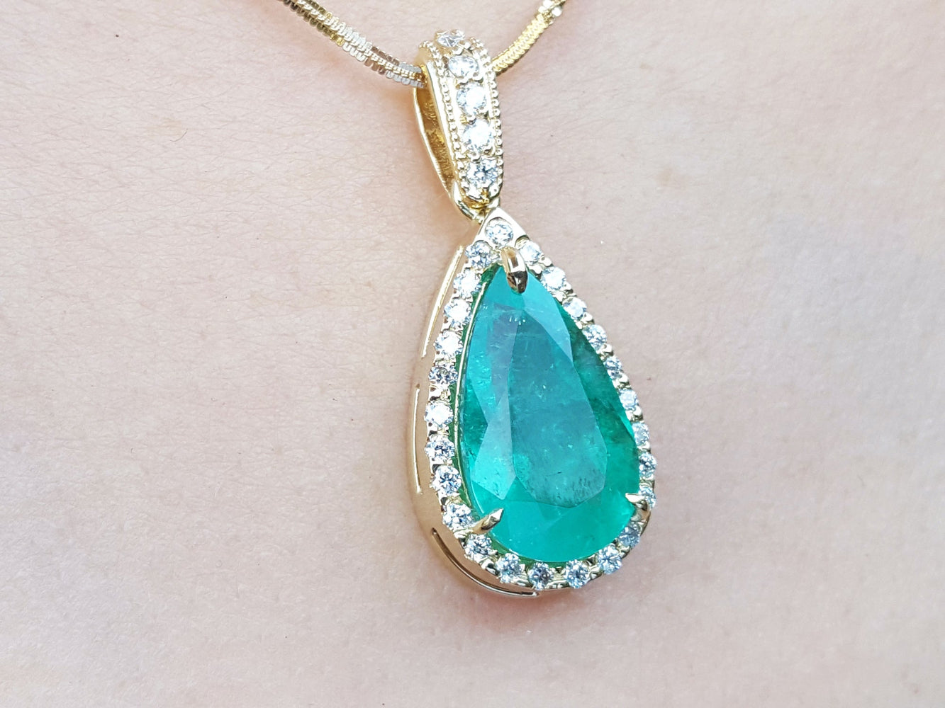 Teardrop-shaped emerald gemstone pendant with halo diamond accents on a light pink skin background