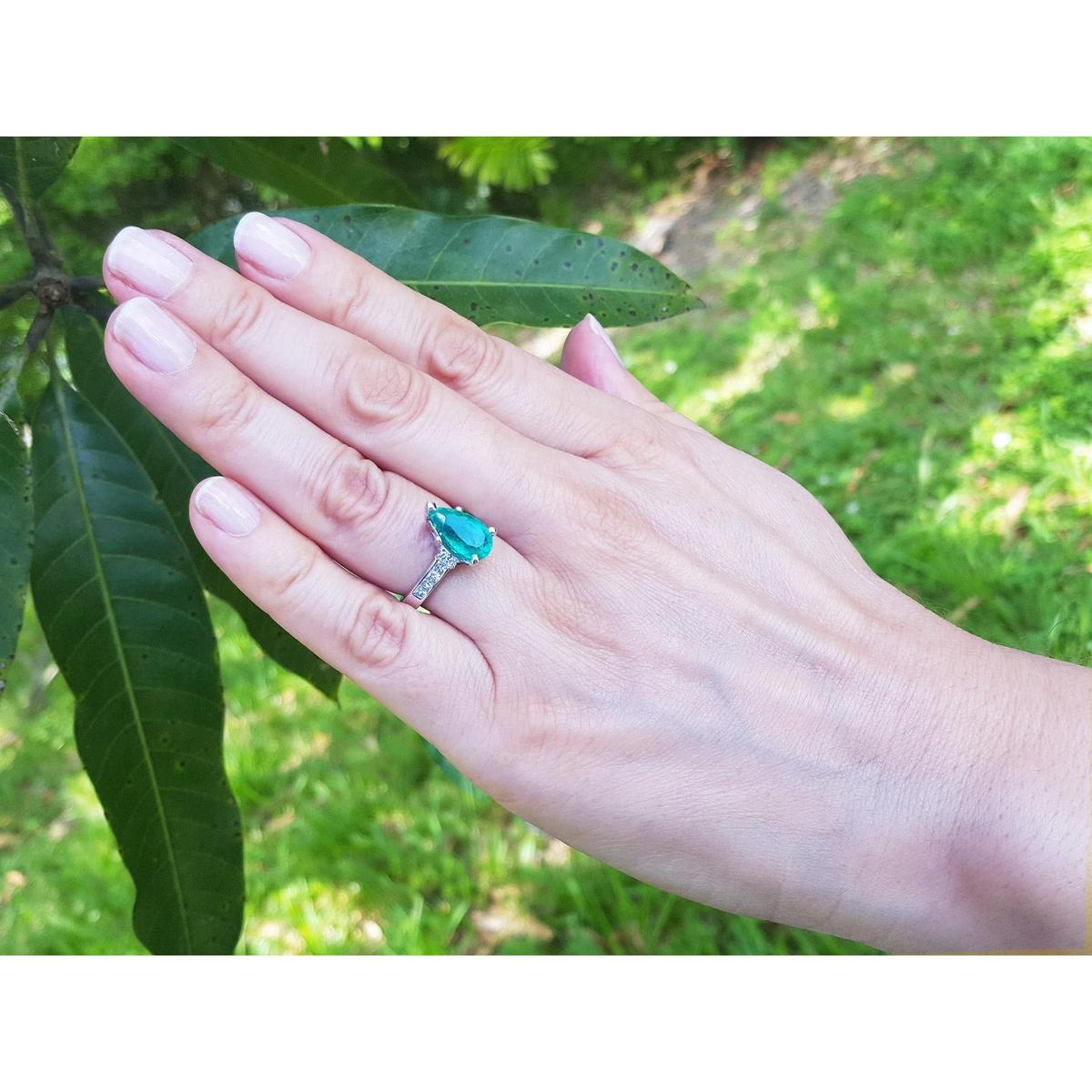 Hand wearing a teardrop emerald ring with greenery in the background.