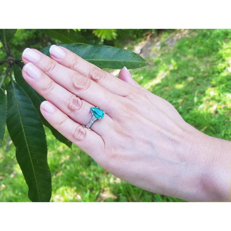 Pear-shaped deep bluish-green emerald ring with princess cut diamonds in 14K white gold on a hand.