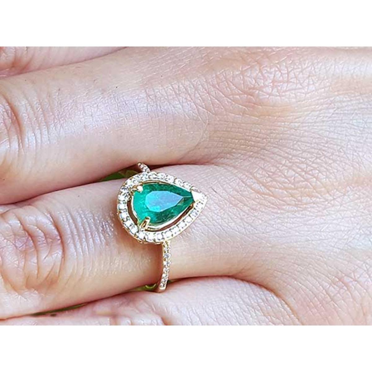 A pear cut emerald encircled by a halo diamond in yellow gold ring on a finger.