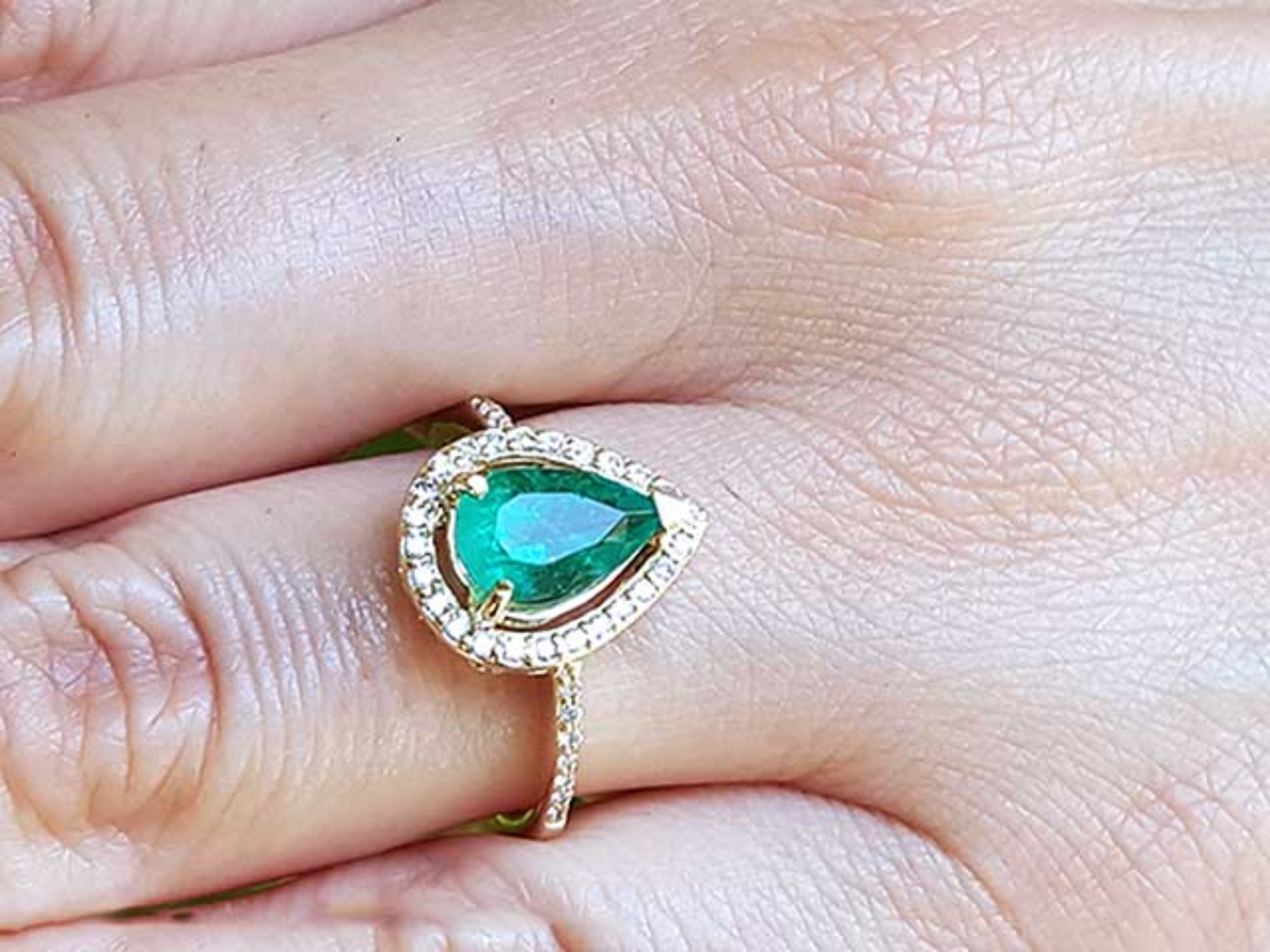 A pear cut emerald encircled by a halo diamond in yellow gold ring on a finger.