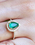 A pear cut emerald encircled by a halo diamond in yellow gold ring on a finger.