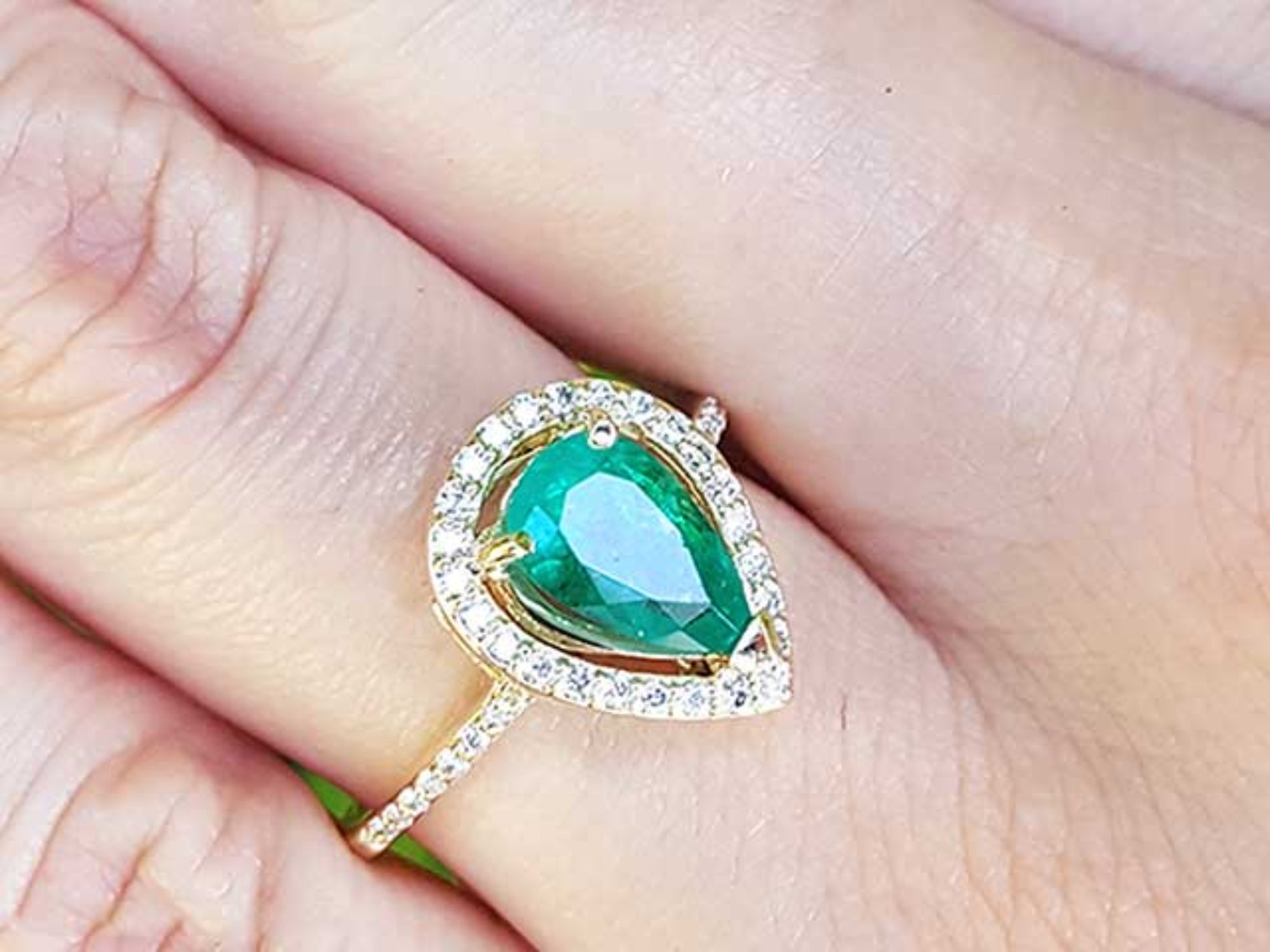 A pear cut emerald in a halo diamond ring on a finger.