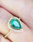 A pear cut emerald in a halo diamond ring on a finger.