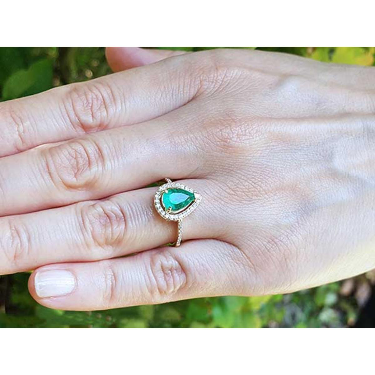 Hand wearing a gold ring with a pear cut emerald into a halo diamond against a blurred natural background.