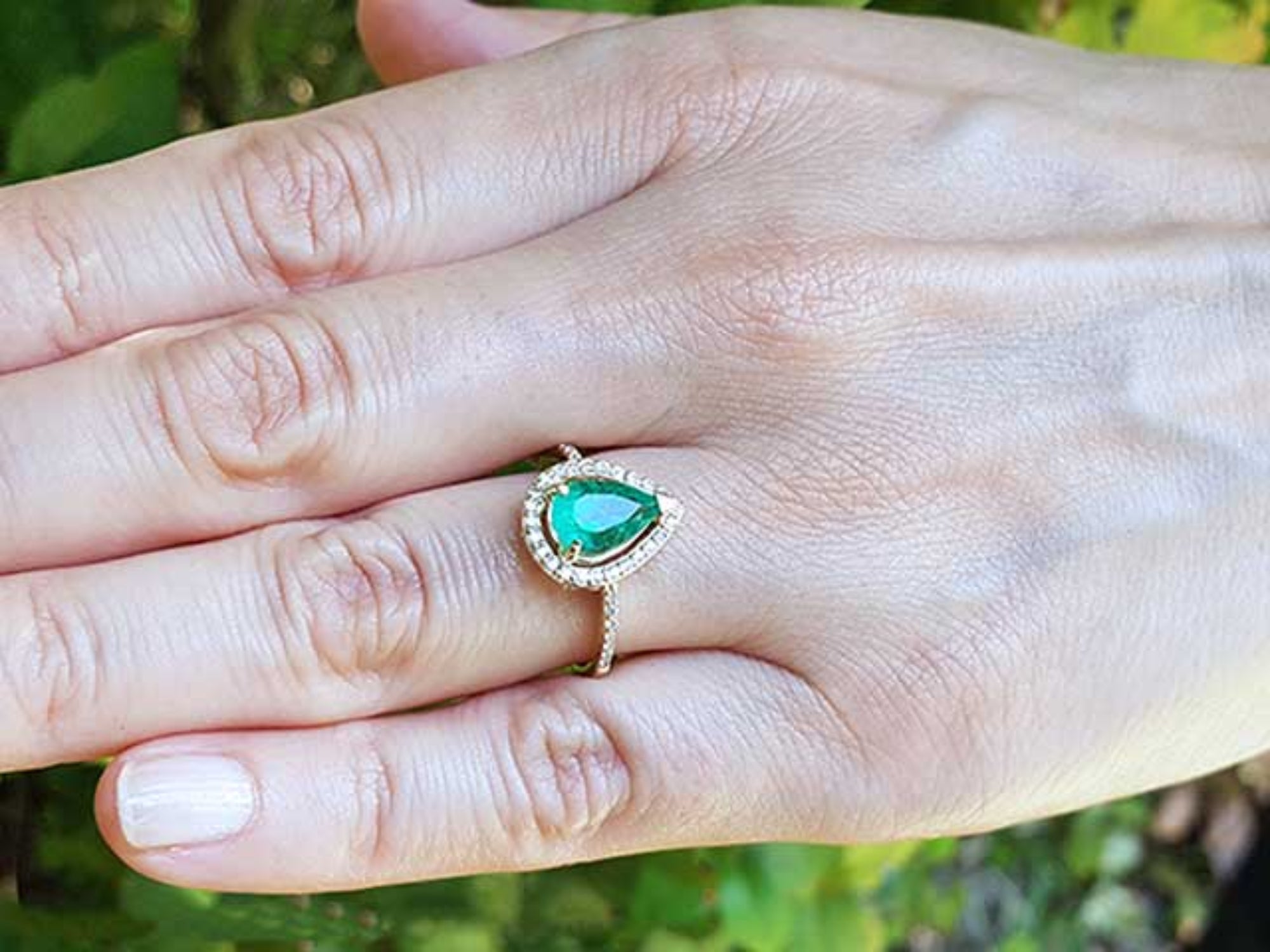 Hand wearing a gold ring with a pear cut emerald into a halo diamond against a blurred natural background.