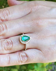 Hand wearing a gold ring with a pear cut emerald into a halo diamond against a blurred natural background.