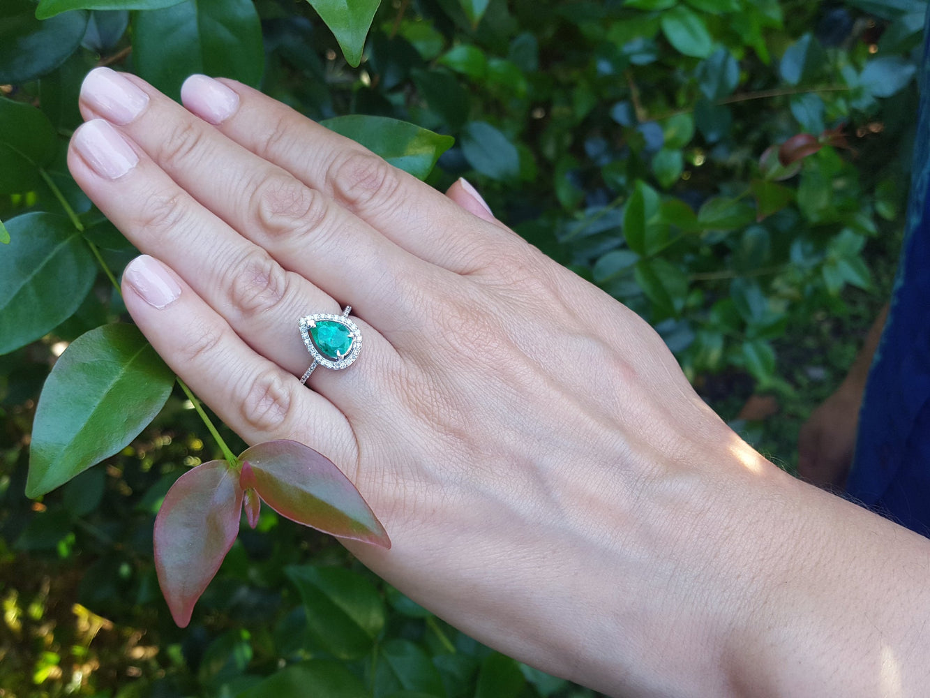 Hand wearing a teardrop emerald ring with a blurred green leafy background.