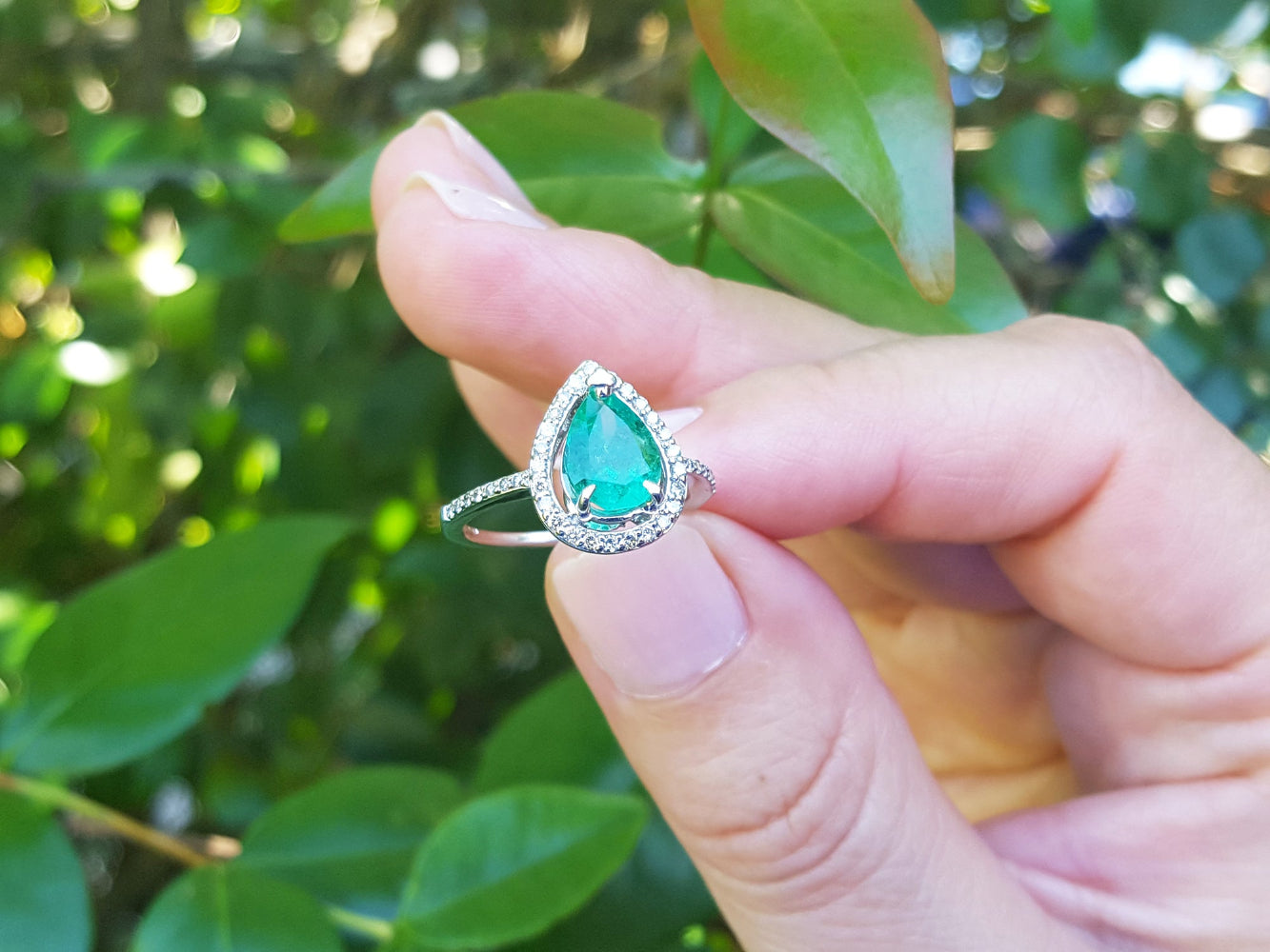 Hand holding between finger a pear cut emerald ring against a blurred green leafy background.