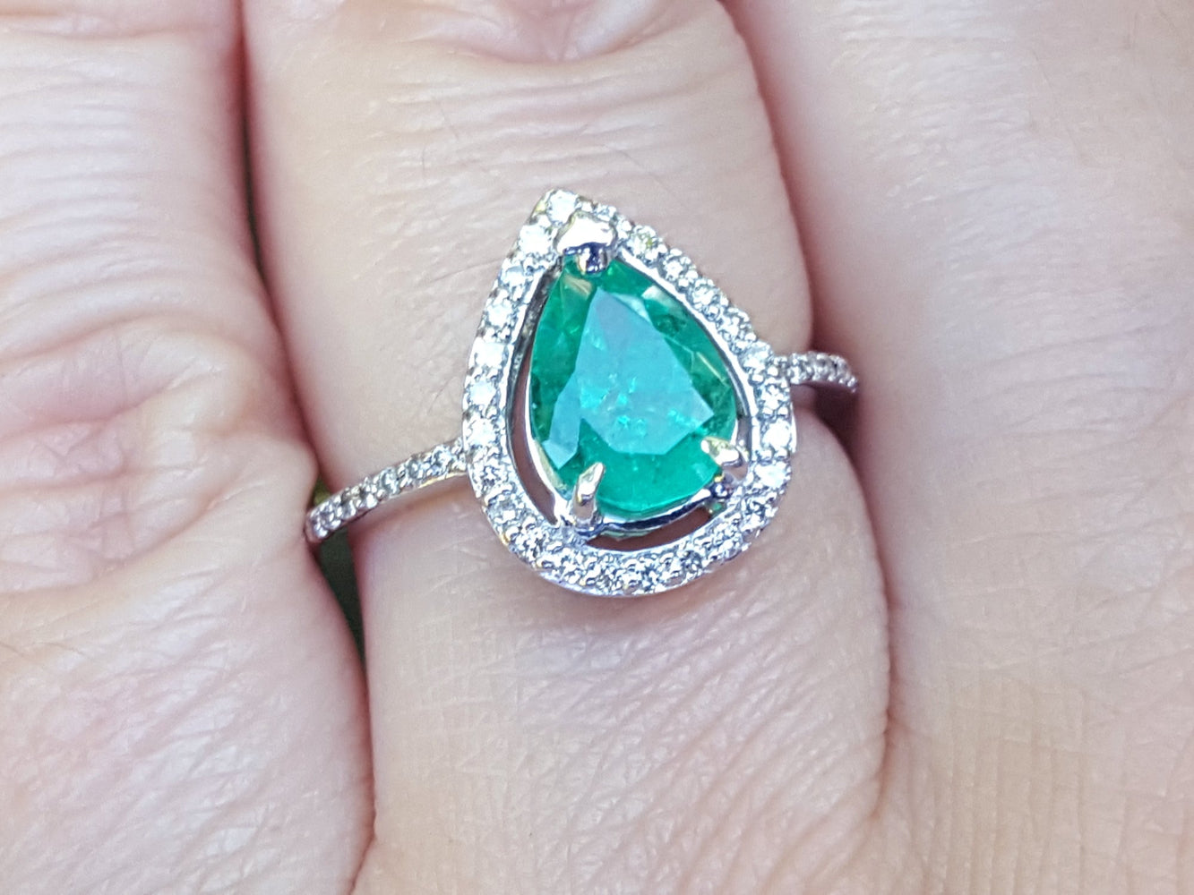 Emerald teardrop ring with diamond halo on a finger.