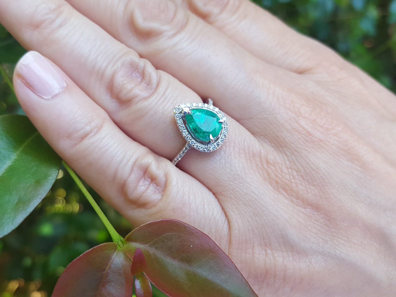 Hand wearing a white gold ring with a pear cut emerald against a natural background.