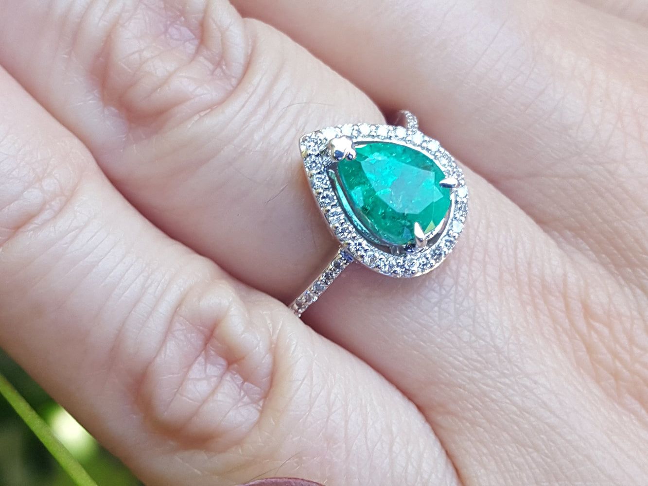 Emerald ring with diamond accents on a finger against a blurred natural background.