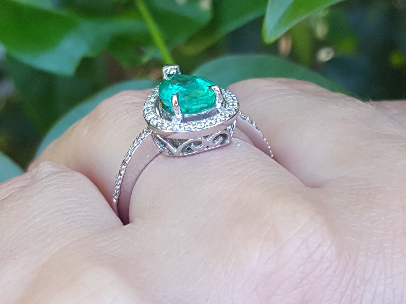 White gold ring with a natural emerald on a hand against a blurred green background.