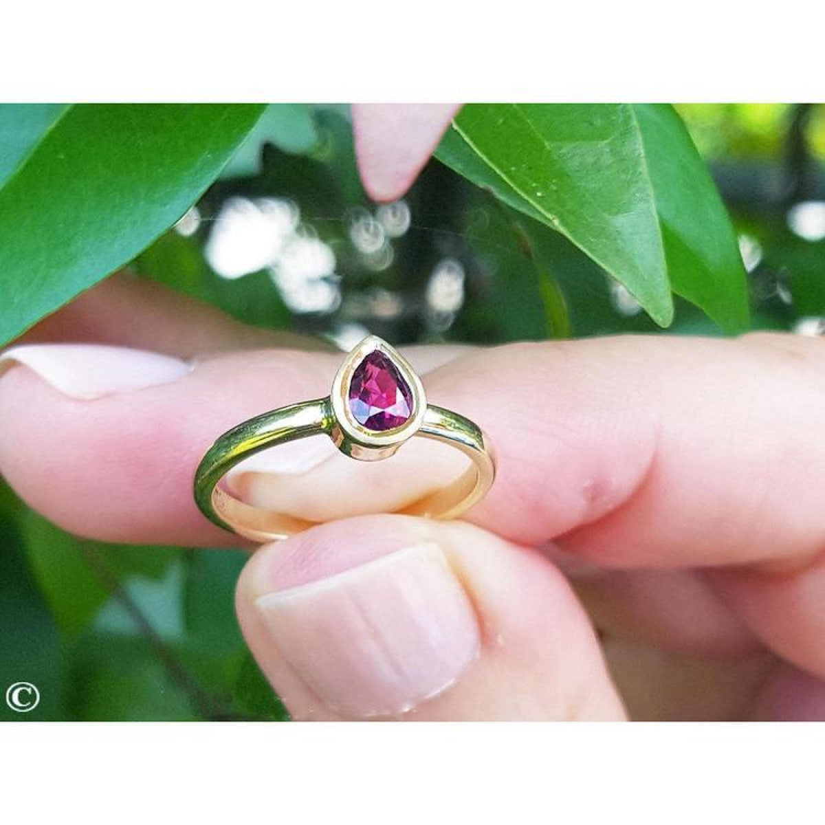 14K yellow gold teardrop natural ruby solitaire ring, held by fingers against green leaves.