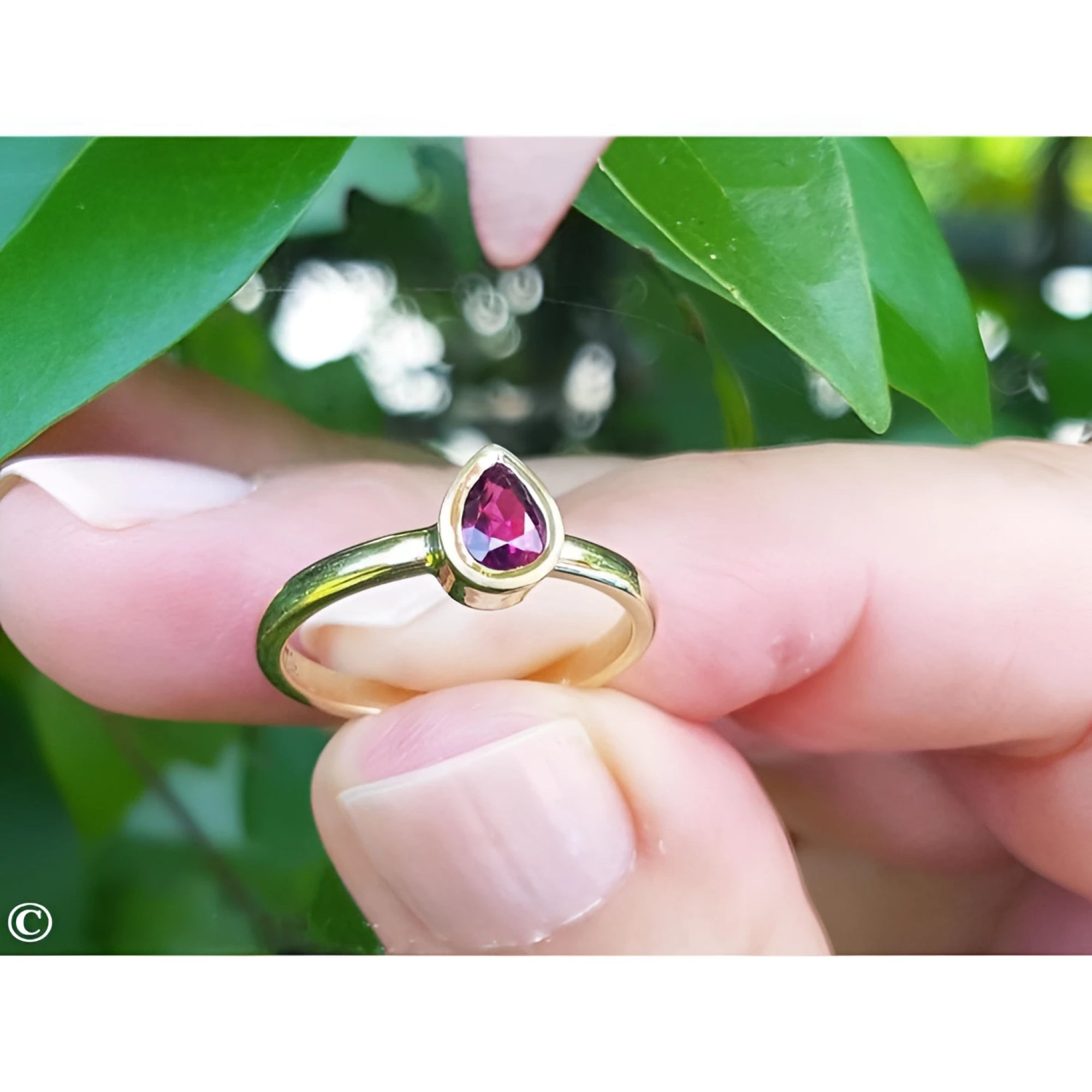 14K yellow gold teardrop natural ruby solitaire ring, held by fingers against green leaves.