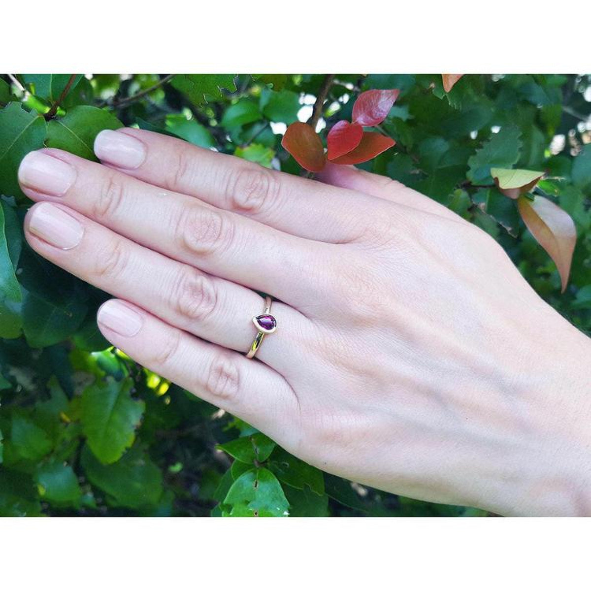 Woman's hand wearing a 14K yellow gold teardrop natural ruby solitaire ring, against green foliage.
