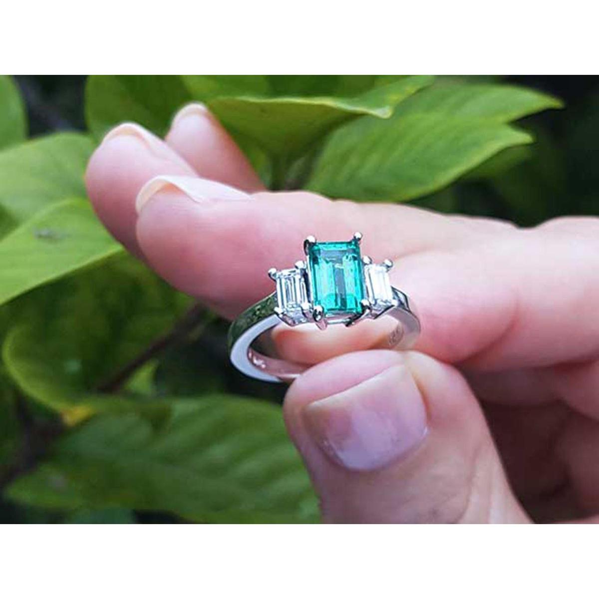 A natural three stone emerald ring for women held in a hand against a green leafy background.