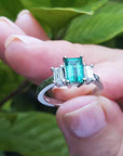 A natural three stone emerald ring for women held in a hand against a green leafy background.