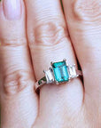 Close-up of a hand wearing an emerald three stone ring against a green leaf background,