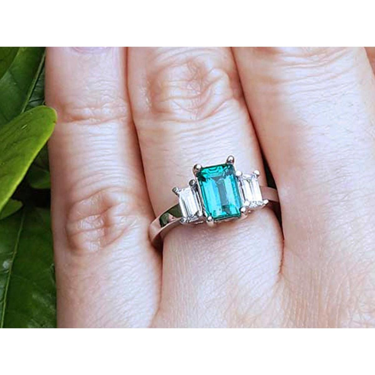 A three stone emerald and side straight baguettes diamond ring on a finger against a natural background.