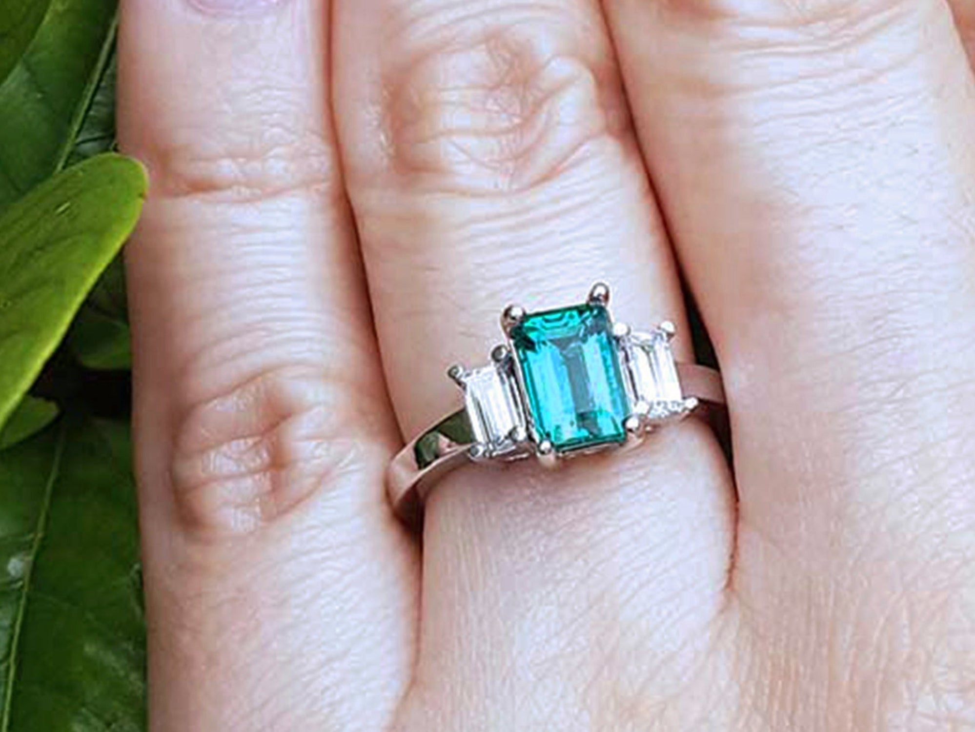 A three stone emerald and side straight baguettes diamond ring on a finger against a natural background.