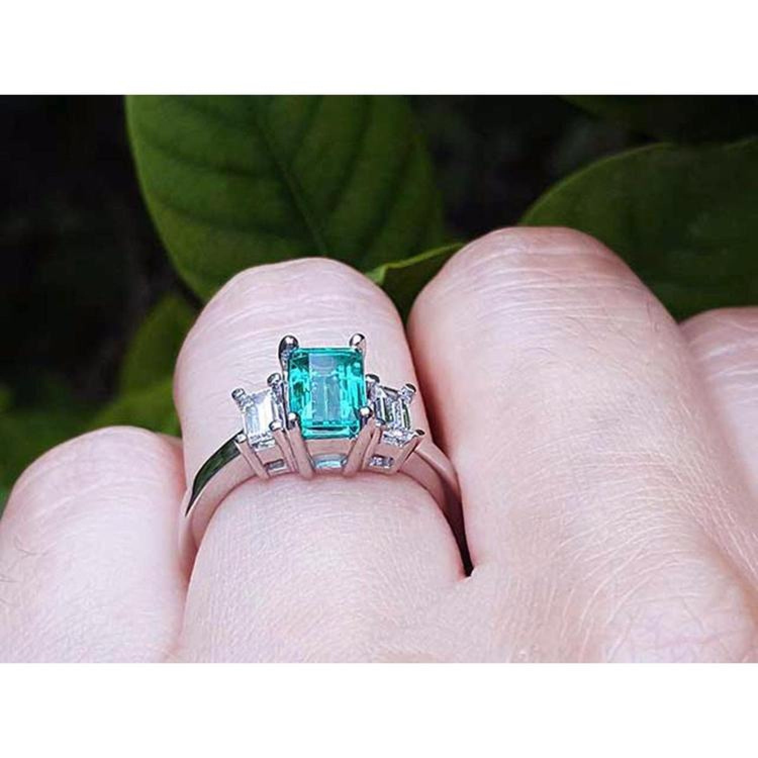 Emerald cut three stone emerald ring with baguette diamonds in 18K white gold on a finger.
