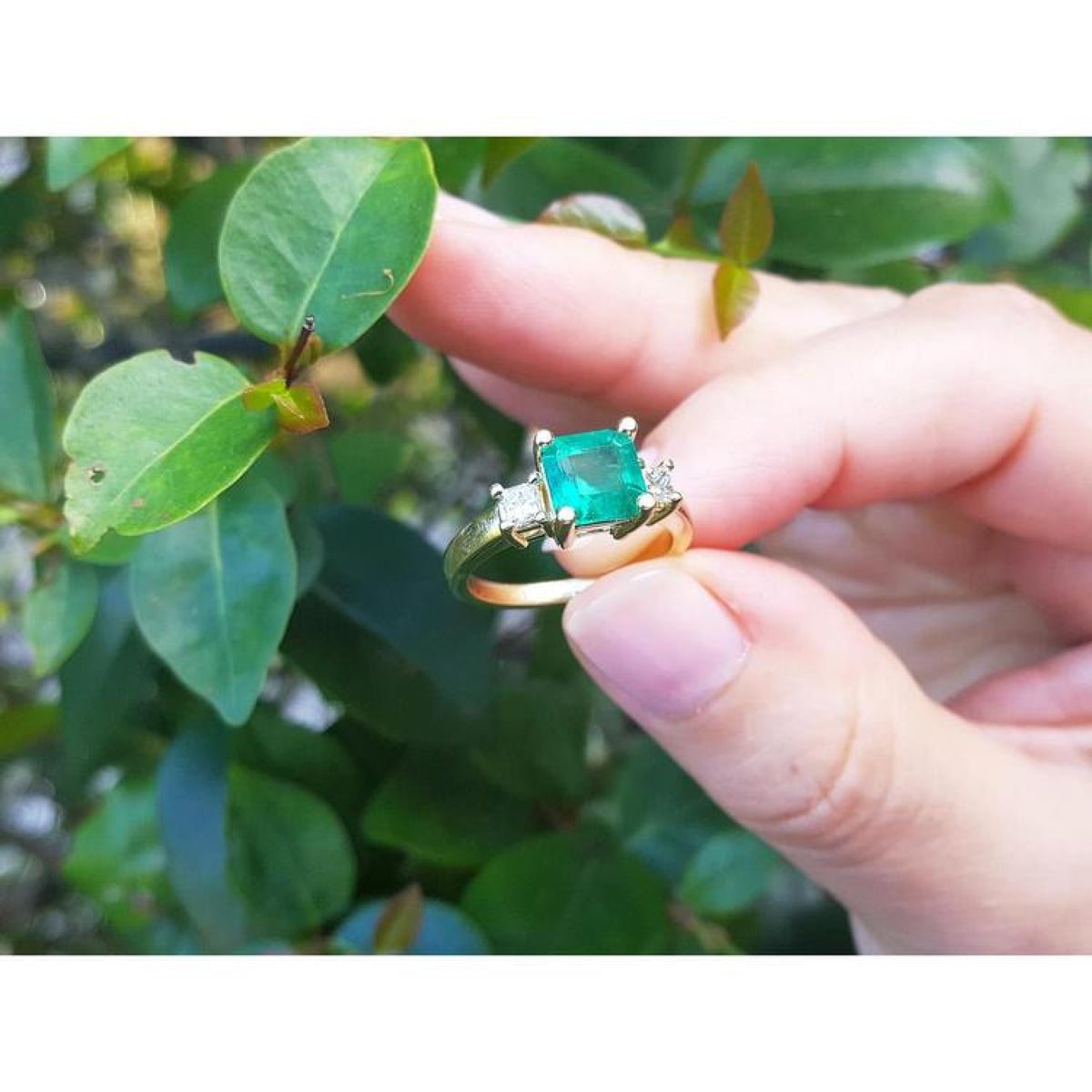 18K yellow gold three-stone ring with a square emerald and princess-cut side diamonds on a hand.