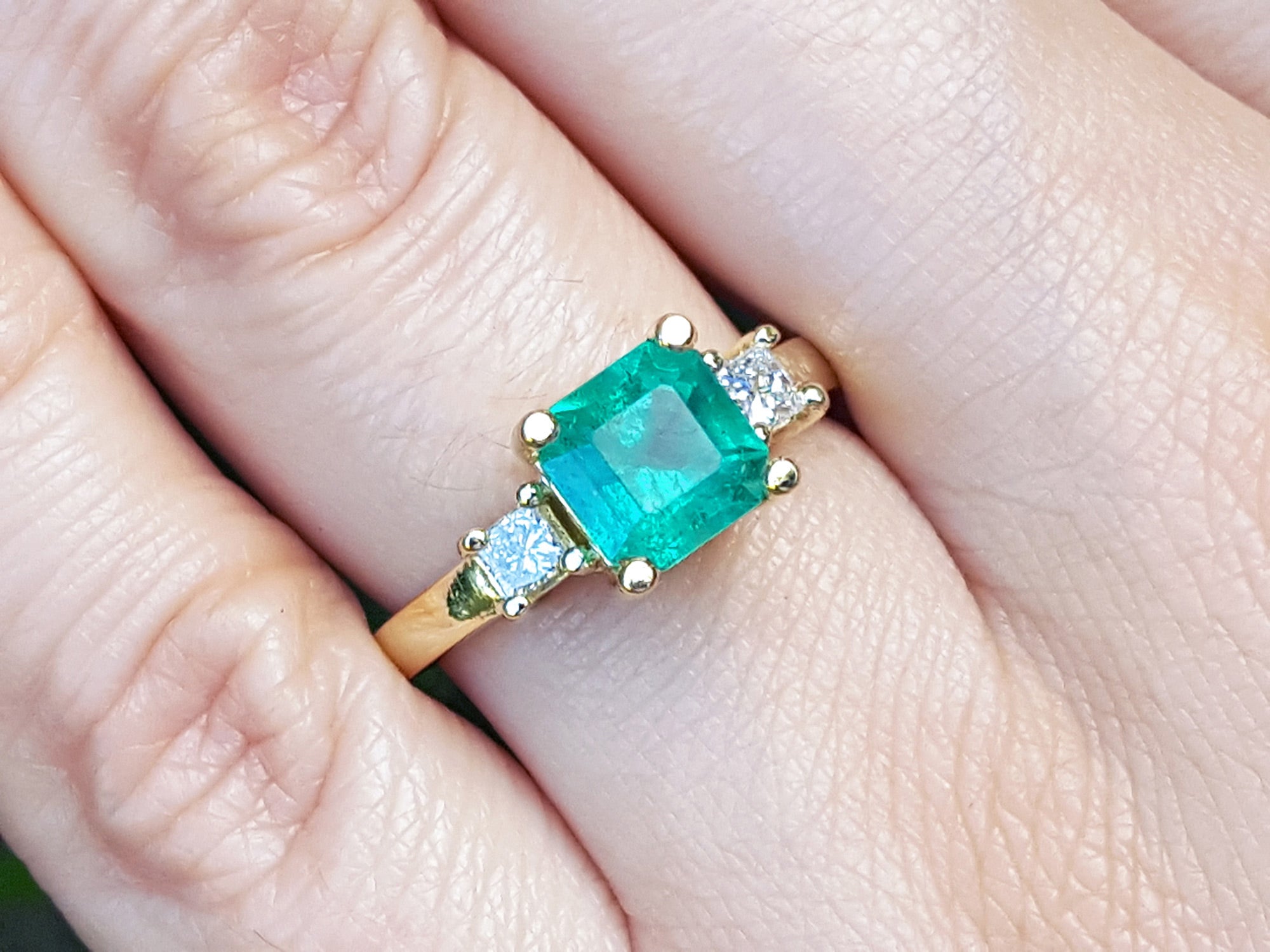 Emerald ring with gold setting and side diamonds on a finger.
