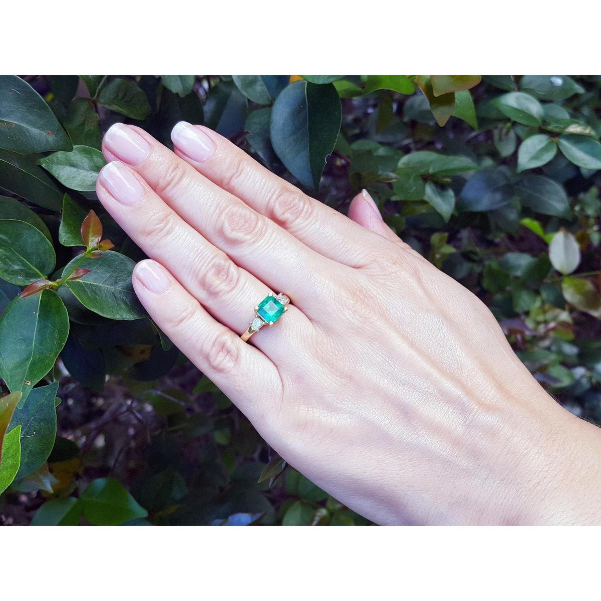 Hand wearing a natural emerald ring with side diamonds against a leafy background.
