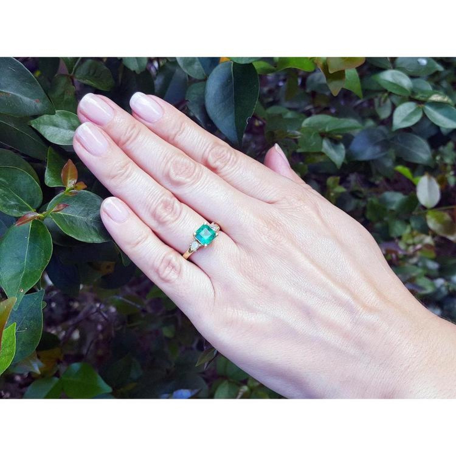 18K yellow gold three-stone ring with a square emerald and princess-cut side diamonds on a hand.