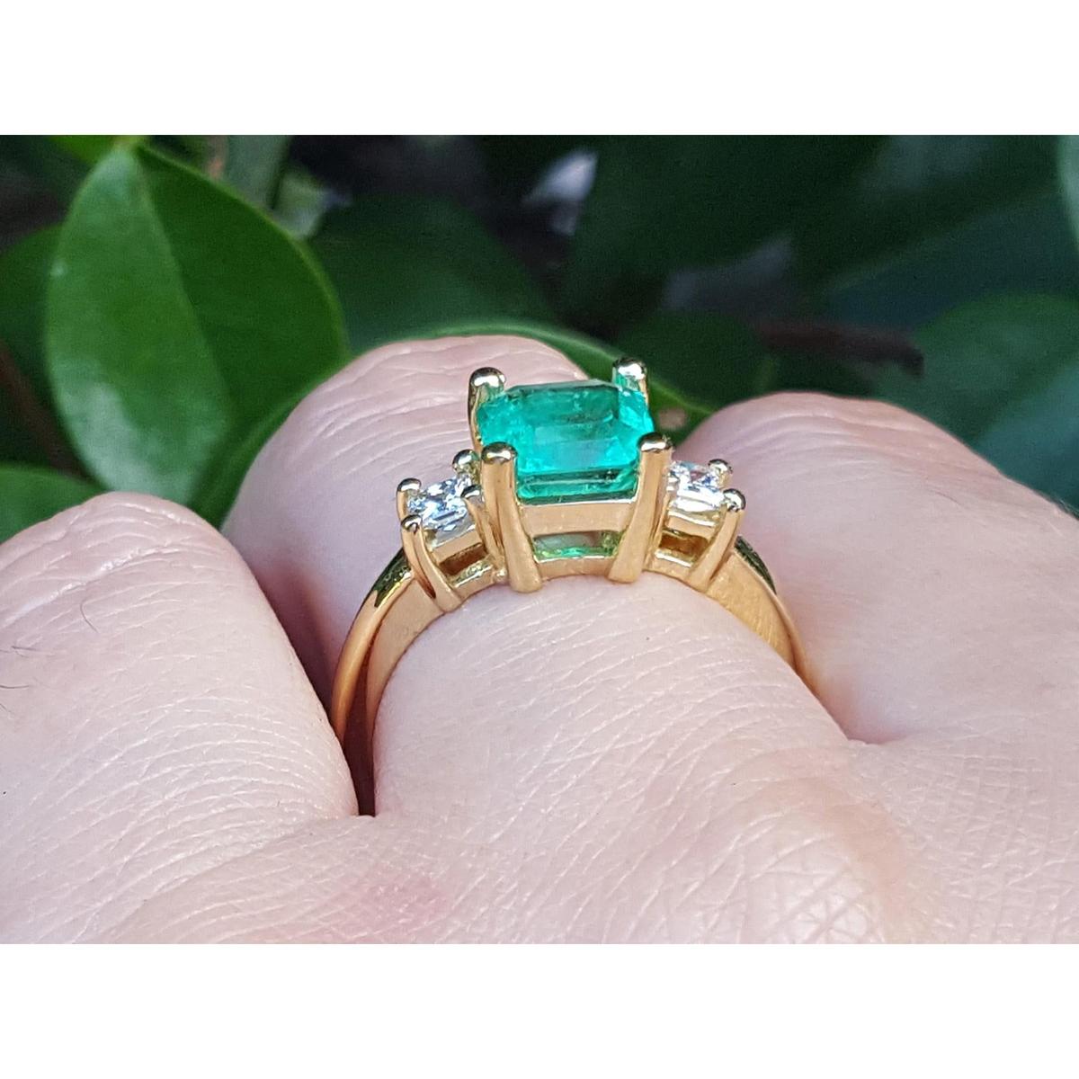 Gold ring with a natural emerald on a finger against a blurred green background.